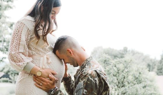 Man in army uniform says goodbye to his pregnant wife, holding and kissing her belly bump.