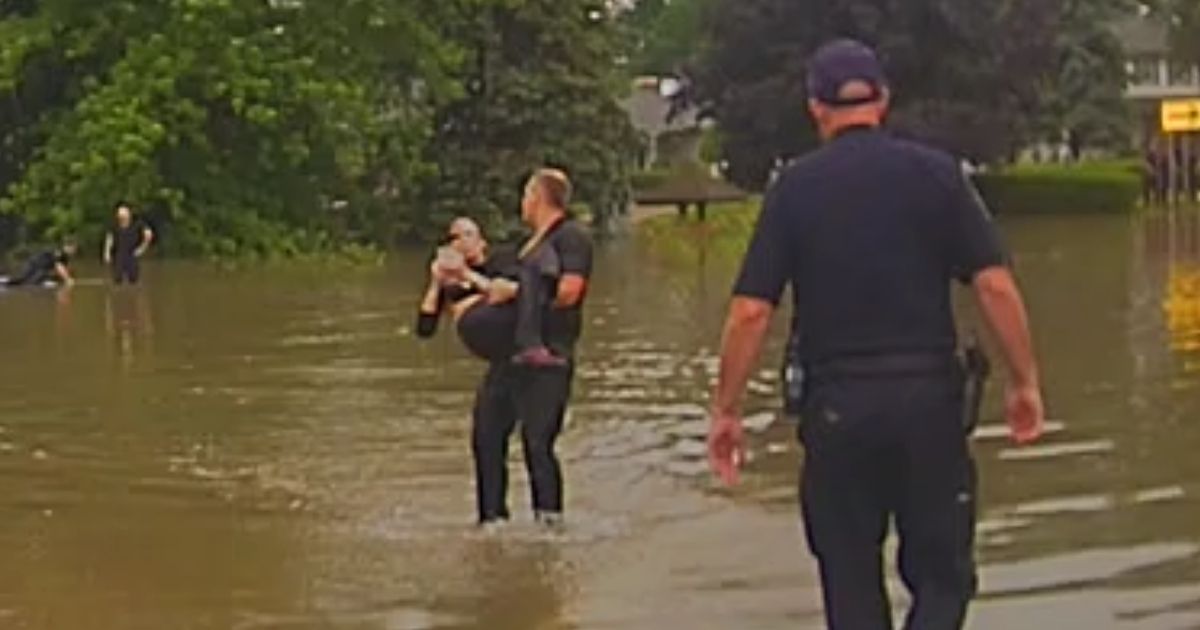 A police officer carries an 82-year-old woman who nearly died in her car after it was swept off the road by floodwaters.