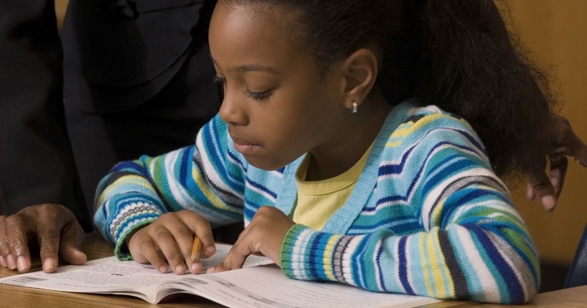 A young female student works in her workbook as a teacher looks over her shoulder.