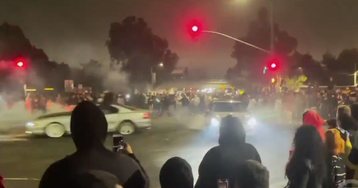 Hundreds of people crowd a traffic light as cars drive through