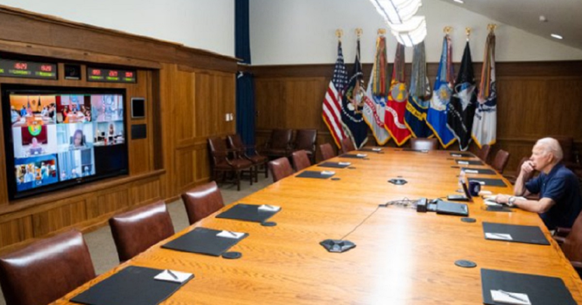 President Joe Biden alone at a conference table watching a video screen across the room.