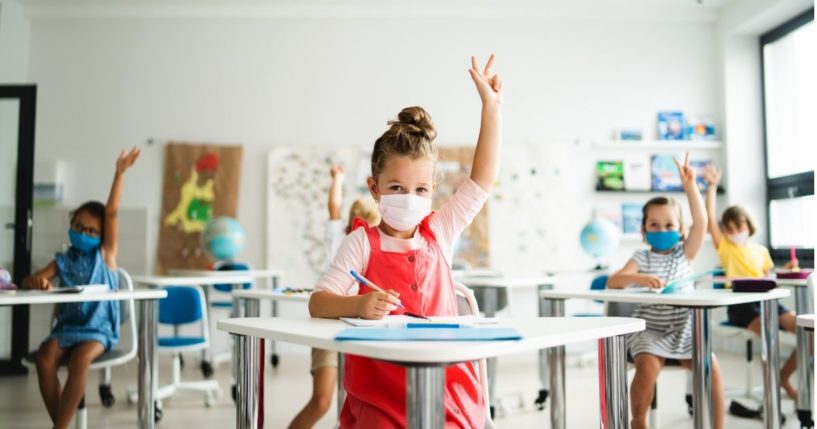 A stock photo shows children with masks sitting at their desks in the classroom, raising their hands.