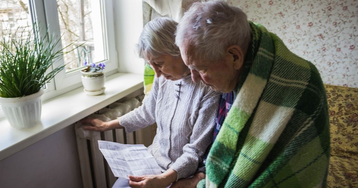 An elderly husband and wife look at their heating bill while sitting in front of a radiator for warmth.