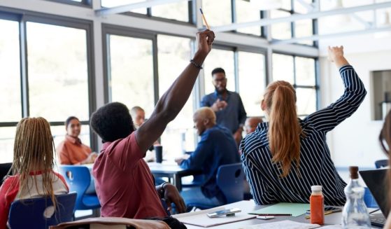 A stock photo shows students at their desks raising their hands while the teacher instructs the class.