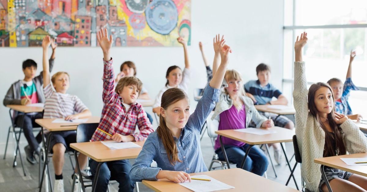 A stock photo shows students sitting in class with their hands raised.