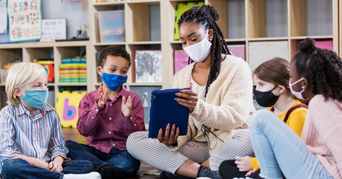 A stock photo shows a group of masked students and their teacher sitting on the floor of a classroom.