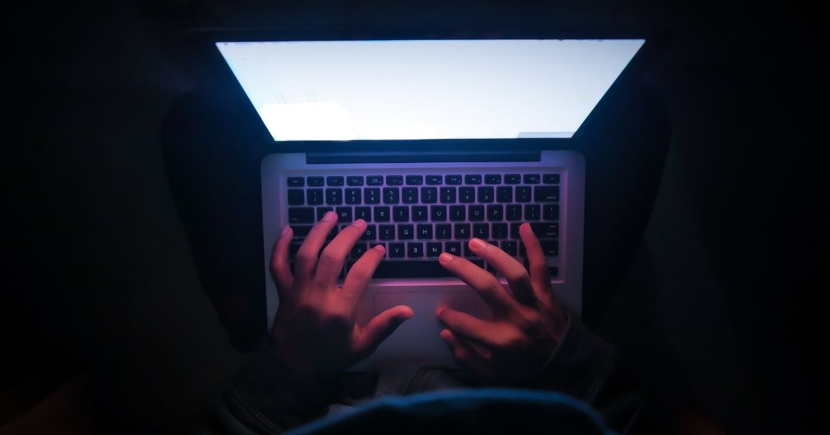 A stock photo shows someone sitting in front of a computer screen in a dark room.