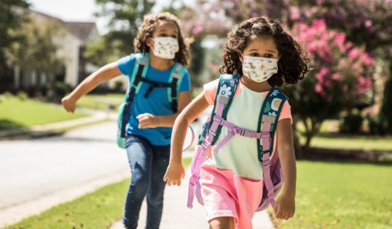 Two children with masks on run outside on a sidewalk