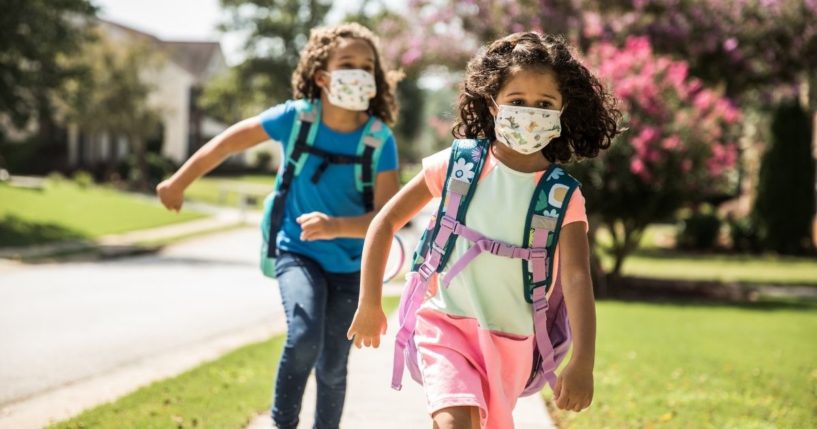 Two children with masks on run outside on a sidewalk