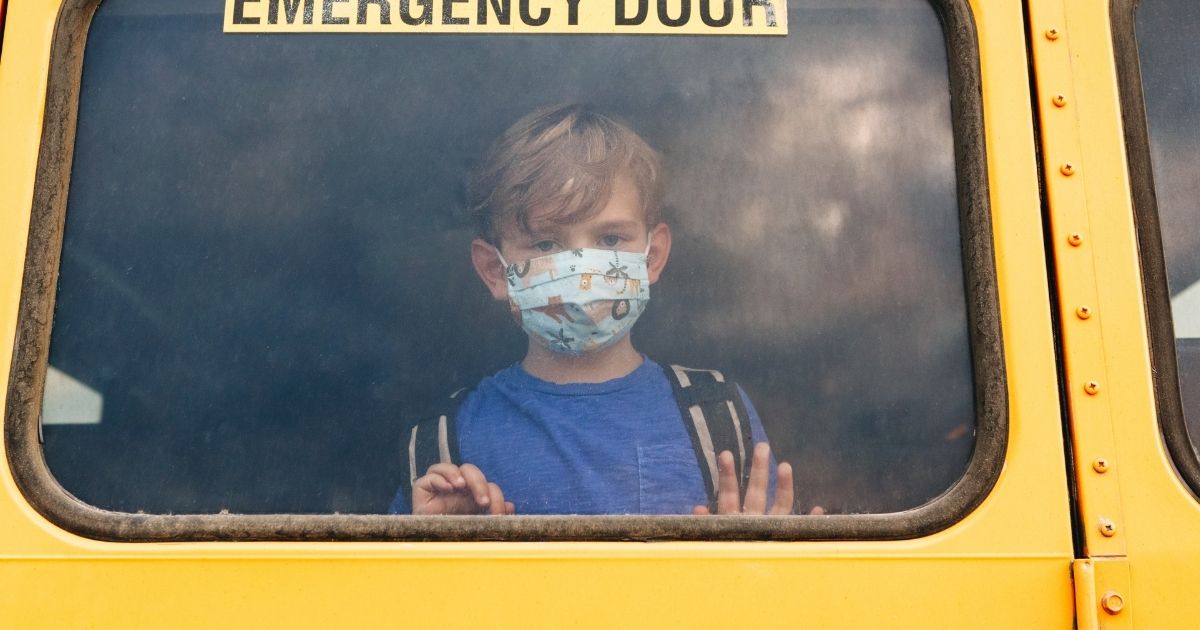 A boy wearing a mask looks out the window of a school bus in this stock image.