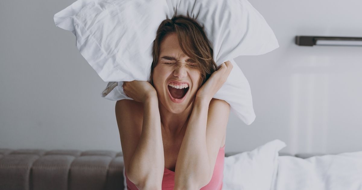 A woman covers her ears with a pillow to deal with an annoying loud noise.