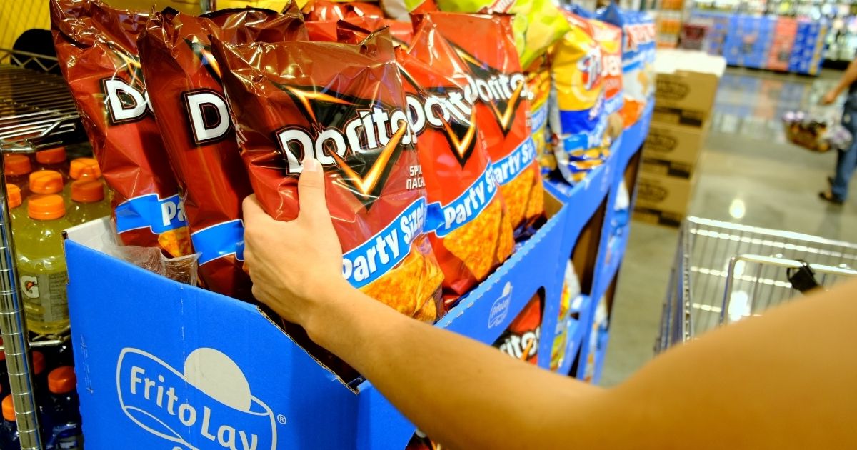 A woman's arm picks up Doritos bag on a supermarket shelf.