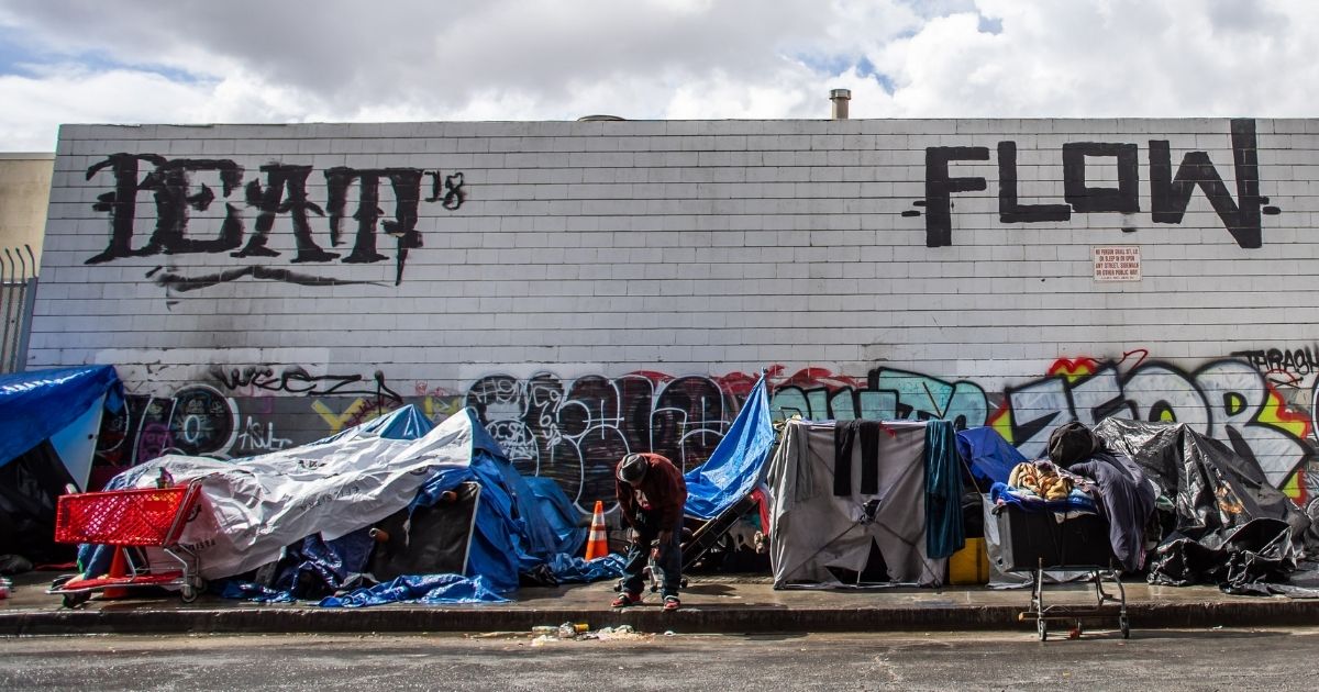 A homeless man stand on 5th Street in Downtown LA, on a sidewalk filled with a makeshift homeless "shanty town."