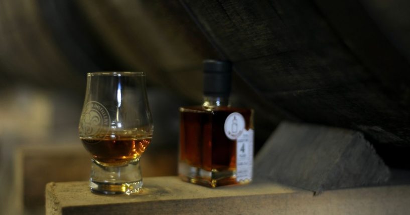 A single malt whiskey sits underneath a wooden barrel.