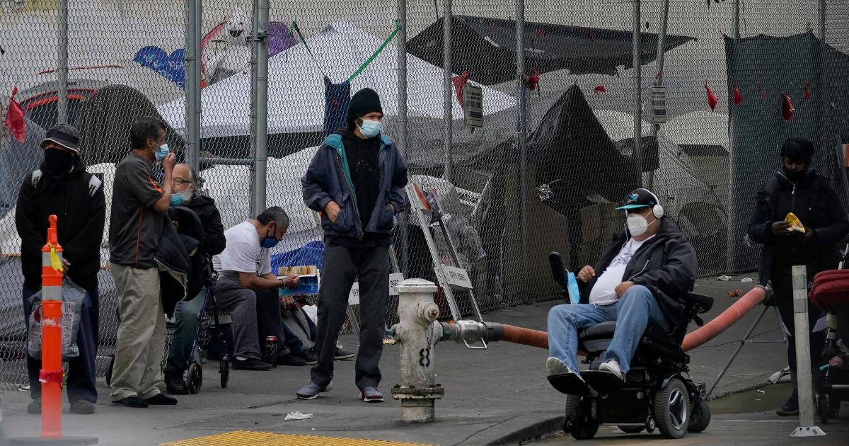 People lingering on a street corner in front of tents set up in a fenced lot in San Francisco