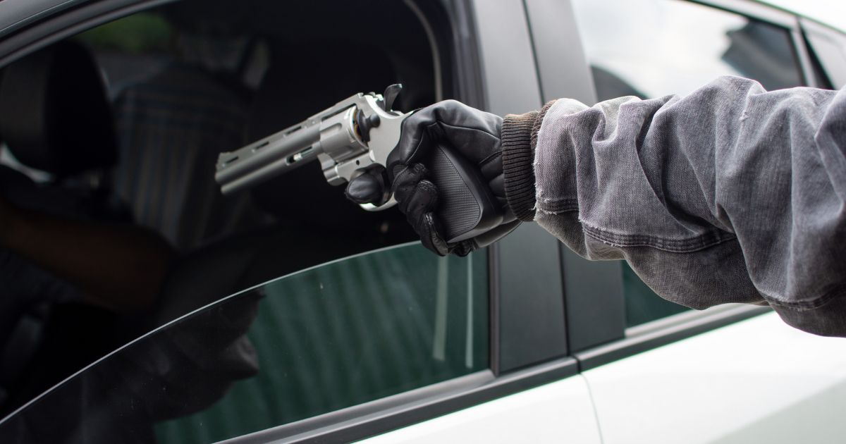 A person pointing a revolver through the open driver-side door of an automobile