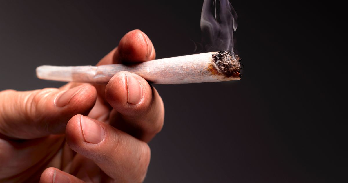 A hand is shown holding a lit marijuana joint.