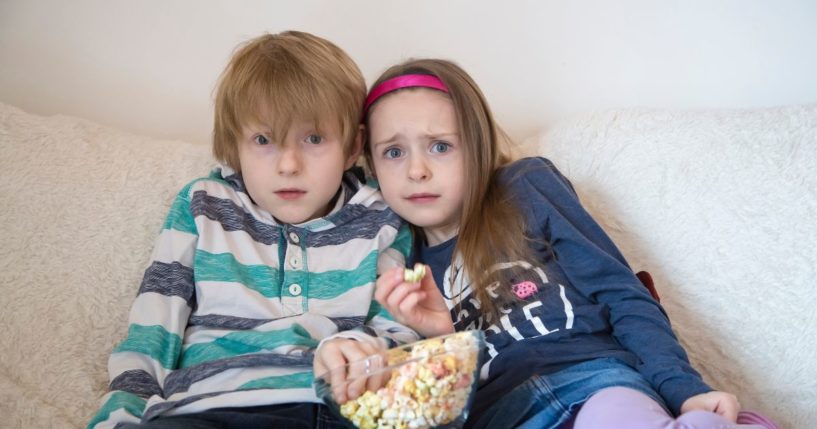 A scared brother and sister watch television with a bowl of colored popcorn.