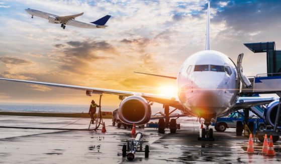 This stock photo shows a passenger plane preparing for flight and getting fueled up at the gate, with another plane taking off in the background.