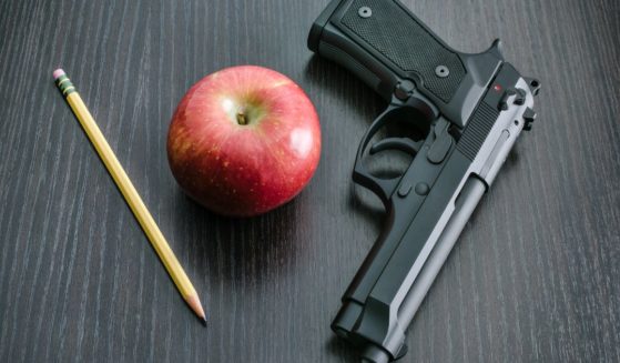 A pencil, an apple, and a handgun are pictured on a teacher's desk.