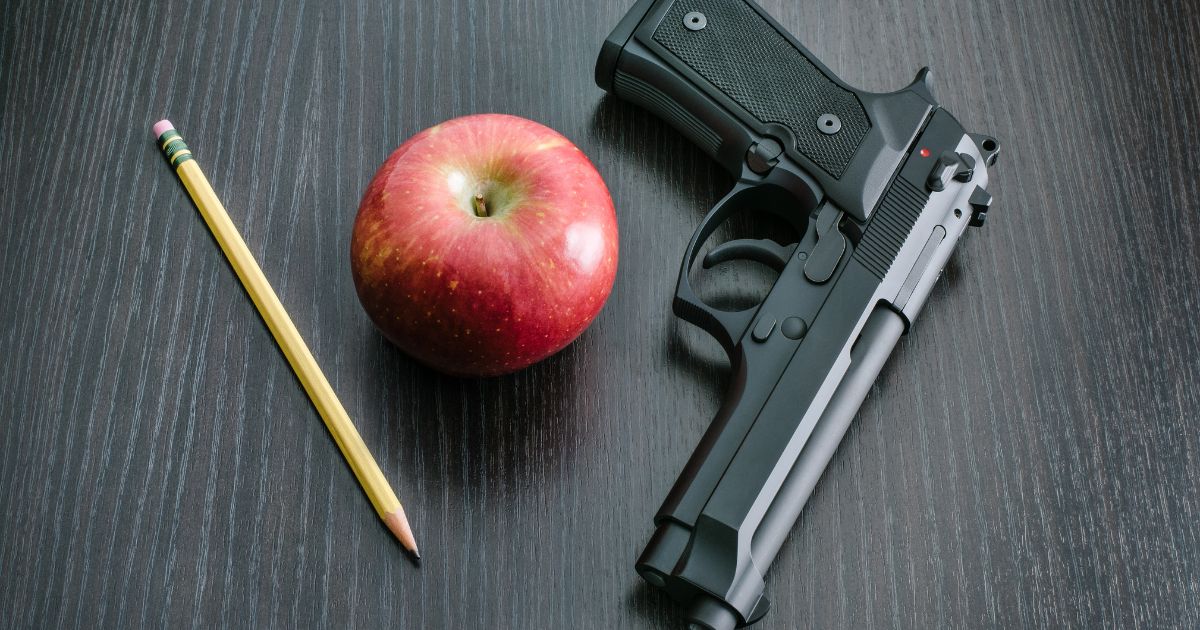 A pencil, an apple, and a handgun are pictured on a teacher's desk.