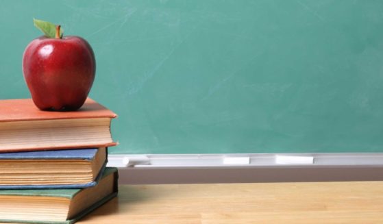 A stock photo shows an apple on a stack of books in a classroom.