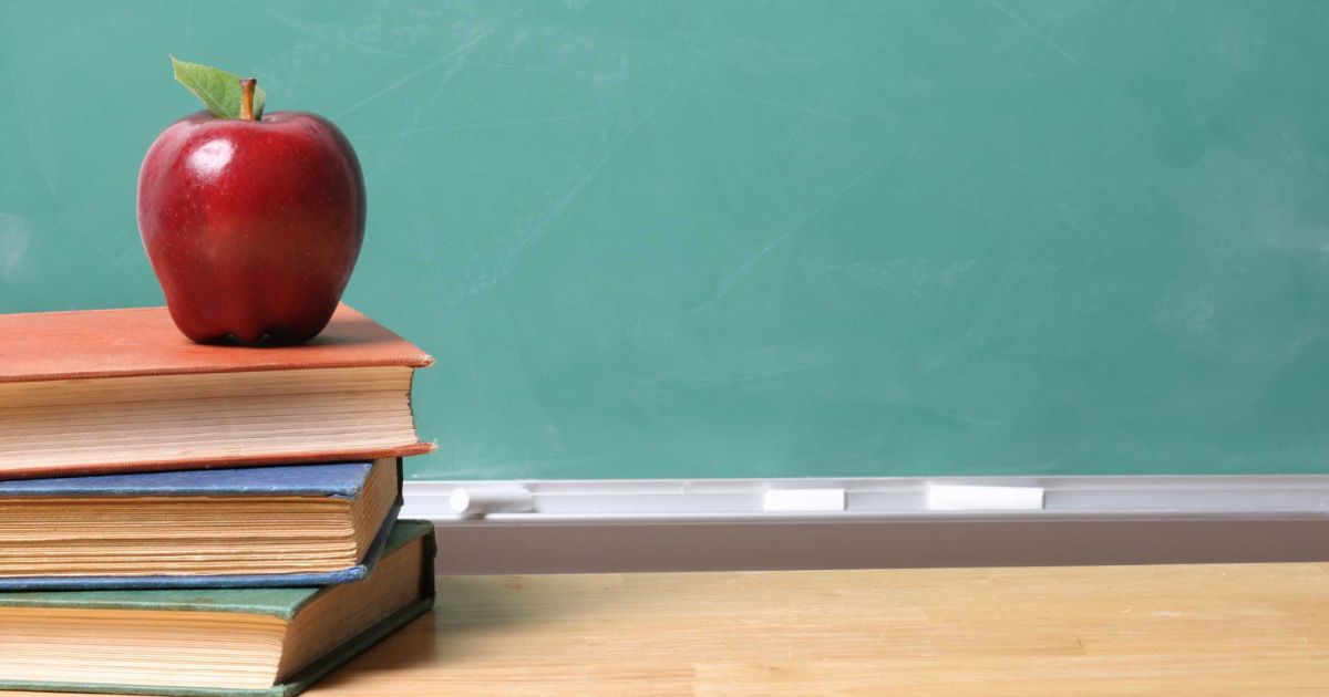 A stock photo shows an apple on a stack of books in a classroom.