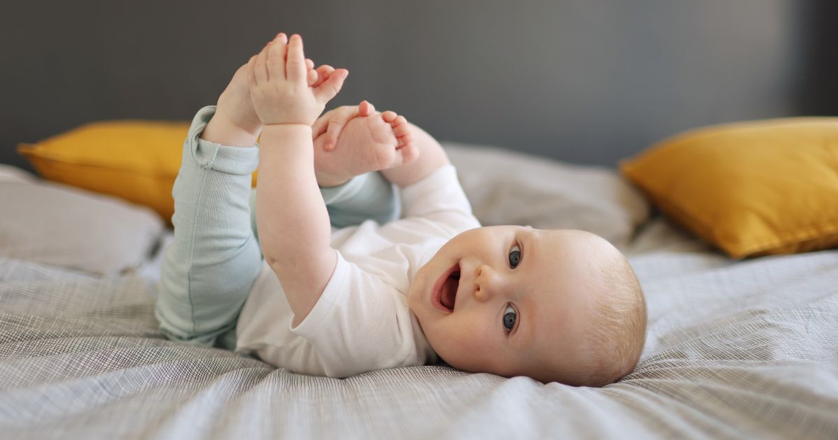 This stock photo shows a 6-month-old baby laying on a bed and smiling.