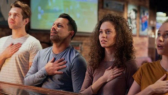 A stock photo shows a group of friends singing the national anthem with their hands over their hearts in a pub.
