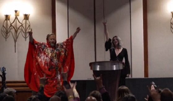 A man in a red caped suit and a woman in black exhort a crowd at a gathering of The Satanic Temple at a Boston hotel.
