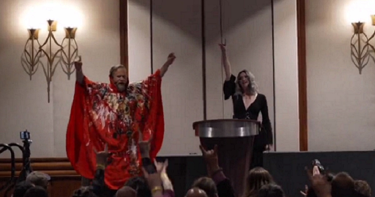 A man in a red caped suit and a woman in black exhort a crowd at a gathering of The Satanic Temple at a Boston hotel.