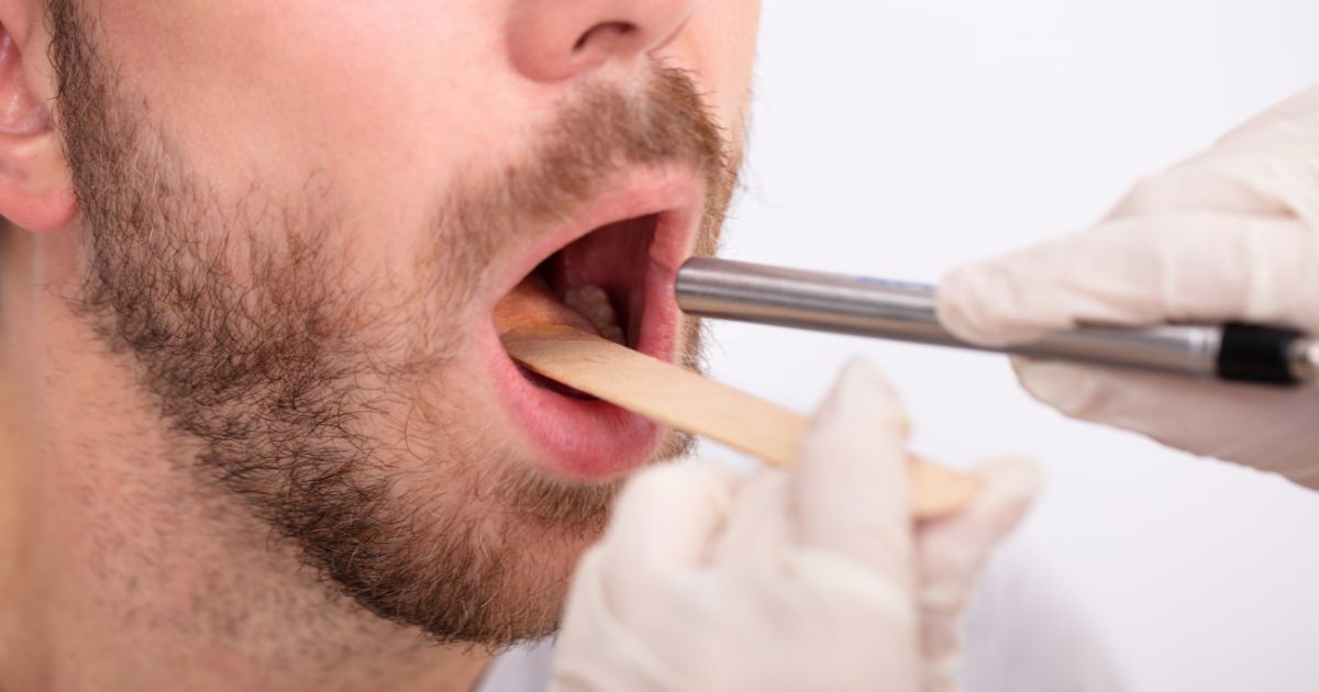 A doctor with a tongue depressor and a flashlight examines a patient's throat in a stock photo.