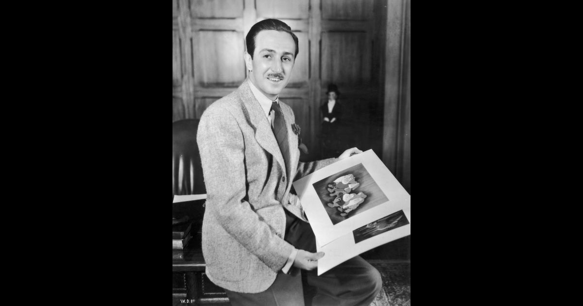Circa 1942: A portrait of American cartoonist and producer Walt Disney (1901 - 1966) seated on the edge of a desk in an office holding illustrations from his animated films 'Snow White and the Seven Dwarfs' and 'Bambi.' Animator David Hand directed both films.