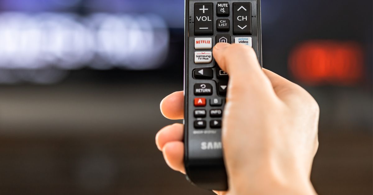 A stock photo shows a hand holding a Samsung television remote control featuring streaming channels such as Netflix and Amazon Prime Video.