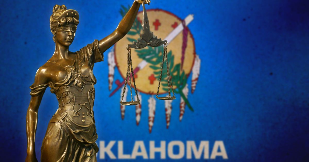A statuette of Lady Justice stands in front of the Oklahoma flag in the above stock image.