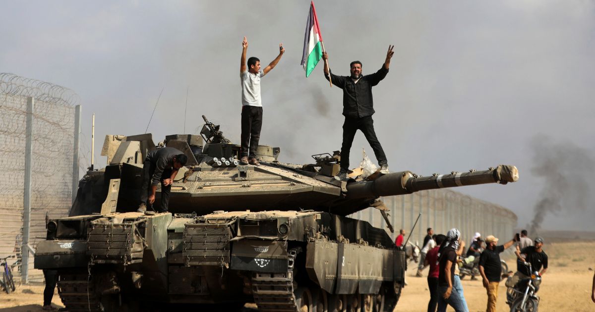 Palestinians waving their flag and celebrating by a destroyed Israeli tank