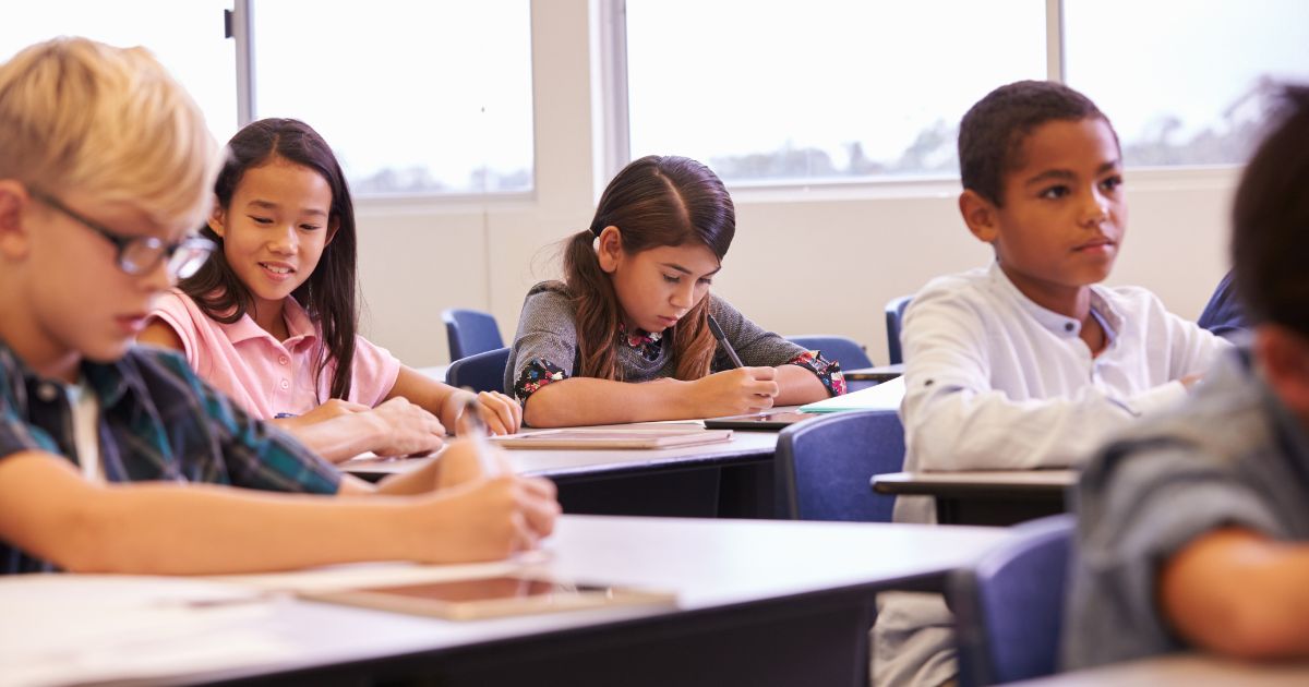 A stock photo shows elementary school kids working at their desks in a classroom.