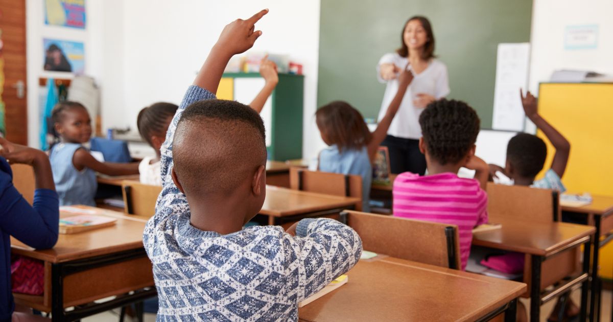 A stock photo shows children raising their hands in a classroom.