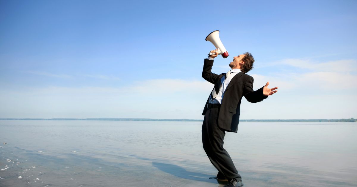 This stock image shows a businessman yelling at the sky with a megaphone.