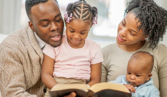 A family of four are sitting on the couch together, reading a Bible story.