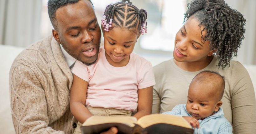 A family of four are sitting on the couch together, reading a Bible story.