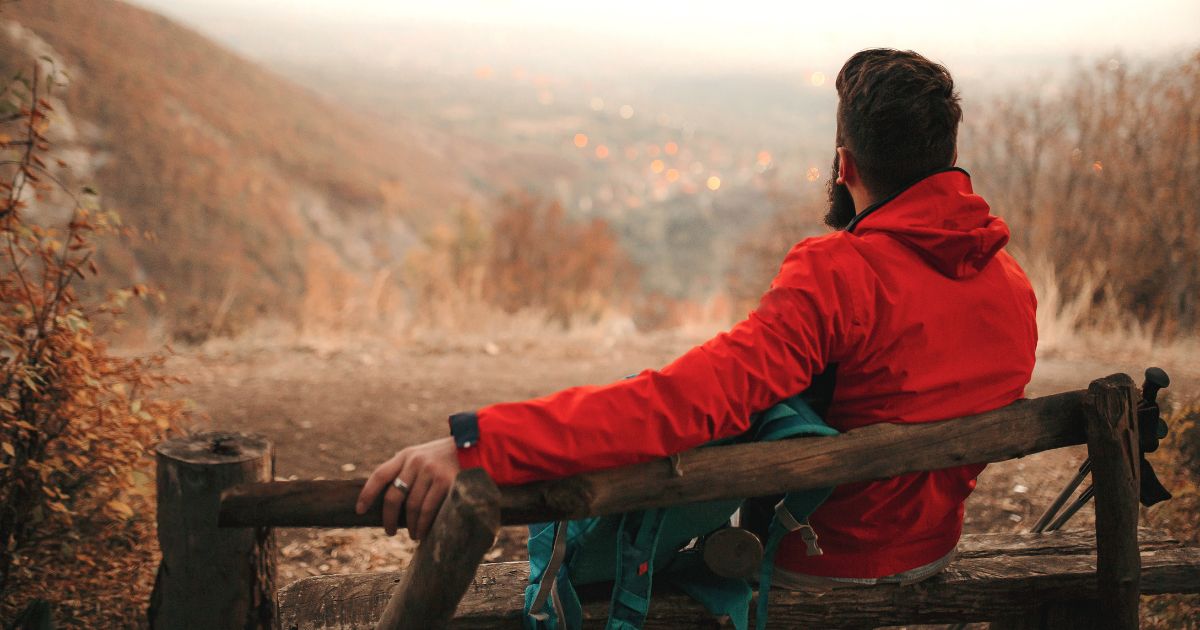 This photo shows a hiker sitting alone on an outdoor bench with his backpack and hiking poles next to him. The spot where he is sitting overlooks city lights in the distance.