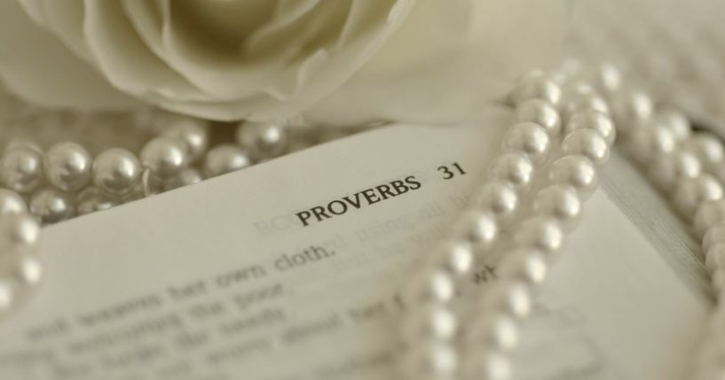 This image shows the Bible opened to the passage of Proverbs 31. A pearl necklace is laying on top of the Bible, and a white rose is laying next the Bible.
