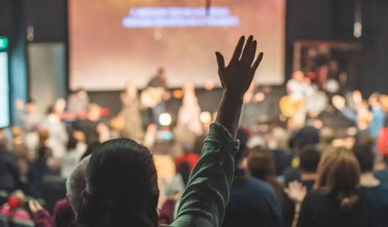 A church congregation worships through music during the service. The back of one woman is in focus, and she is raising a hand in praise.