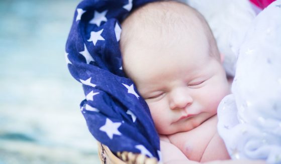 This image shows a newborn baby sleeping in a basket filled with patriotic scarves.