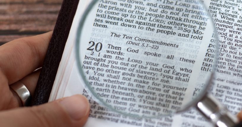 A man's hand holds a magnifying glass over a Bible chapter heading for "The Ten Commandments."