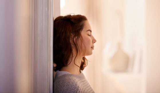 This image shows a young woman leaning her back against a wall while shutting her eyes.