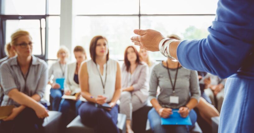 The hand of a trainer addressing a group of females sitting in a conference hall.