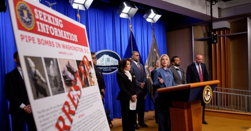 A poster reads "ARRESTED" in bold letters as Attorney General Pam Bondi speaks during a news conference on an arrest of a suspect in the January 6th pipe bomb case at the Department of Justice on Dec. 4, 2025, in Washington, D.C.