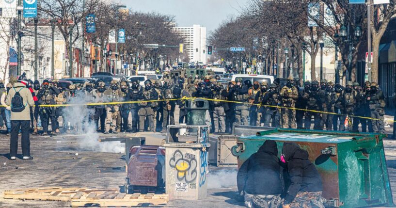 Protesters take cover behind overturned trash containers as a line of federal agents forms a barricade across a downtown street during clashes following the fatal shooting of a demonstrator earlier in the day, on Jan. 24, 2026, in Minneapolis, Minnesota.
